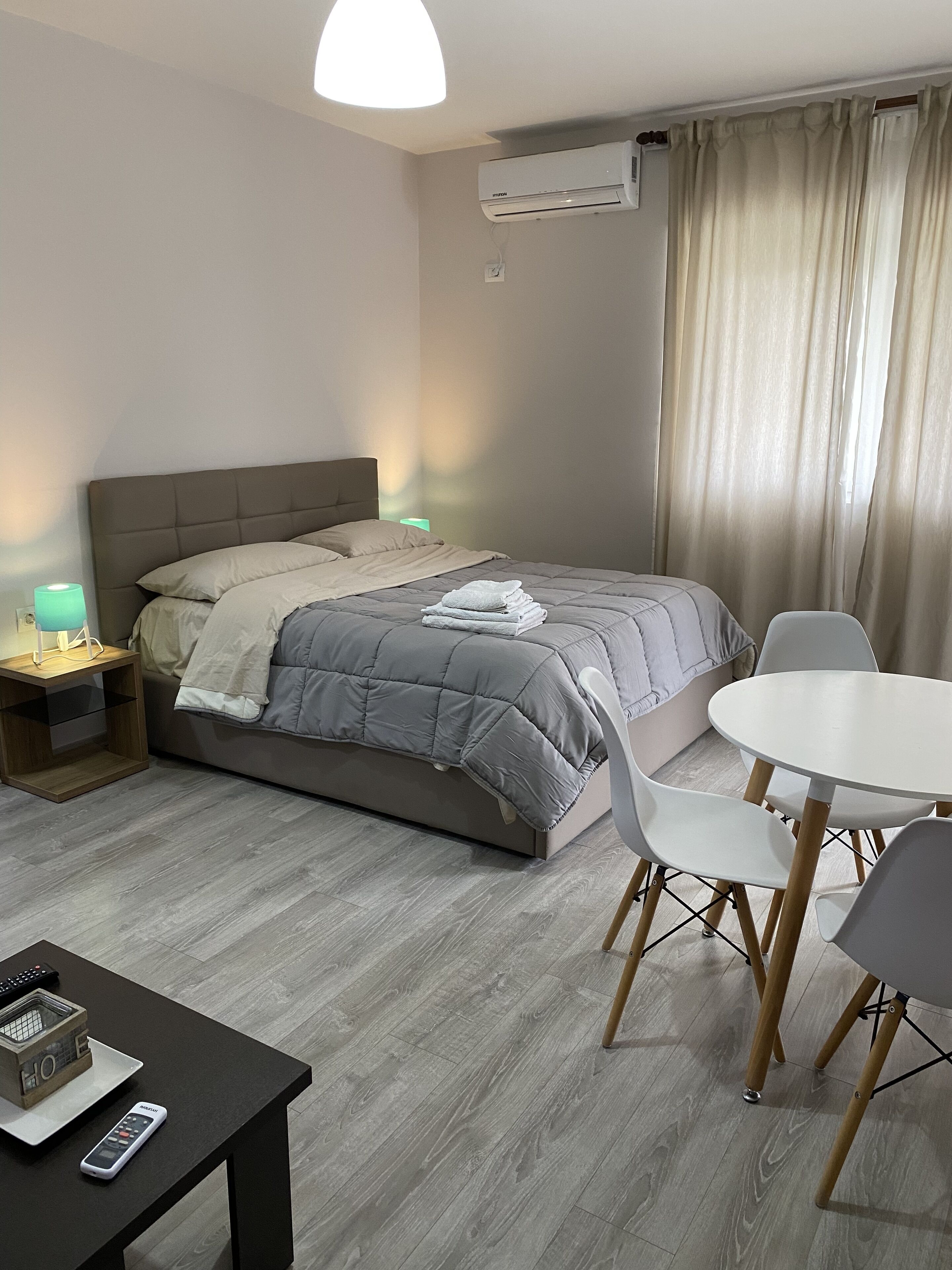 A hotel room with a bed, bedside table, lamp, air conditioning unit, curtains, and a dining area with chairs and a table.