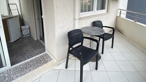 Un petit balcon avec une table et deux chaises, un réfrigérateur et une fenêtre équipée d’un appareil de climatisation.