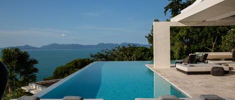 Una piscina d'hidromassatge moderna d'ulls de gat amb una vista clara del mar i les muntanyes, envoltada de gandules i una zona de seients coberta.