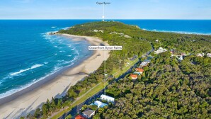 An aerial view of Oceanfront Byron Palms, featuring a beach, a lighthouse, and a residential area with houses and greenery. - Oceanfront Byron Palms - 2 BR Main Beach apartment (Byron Bay)