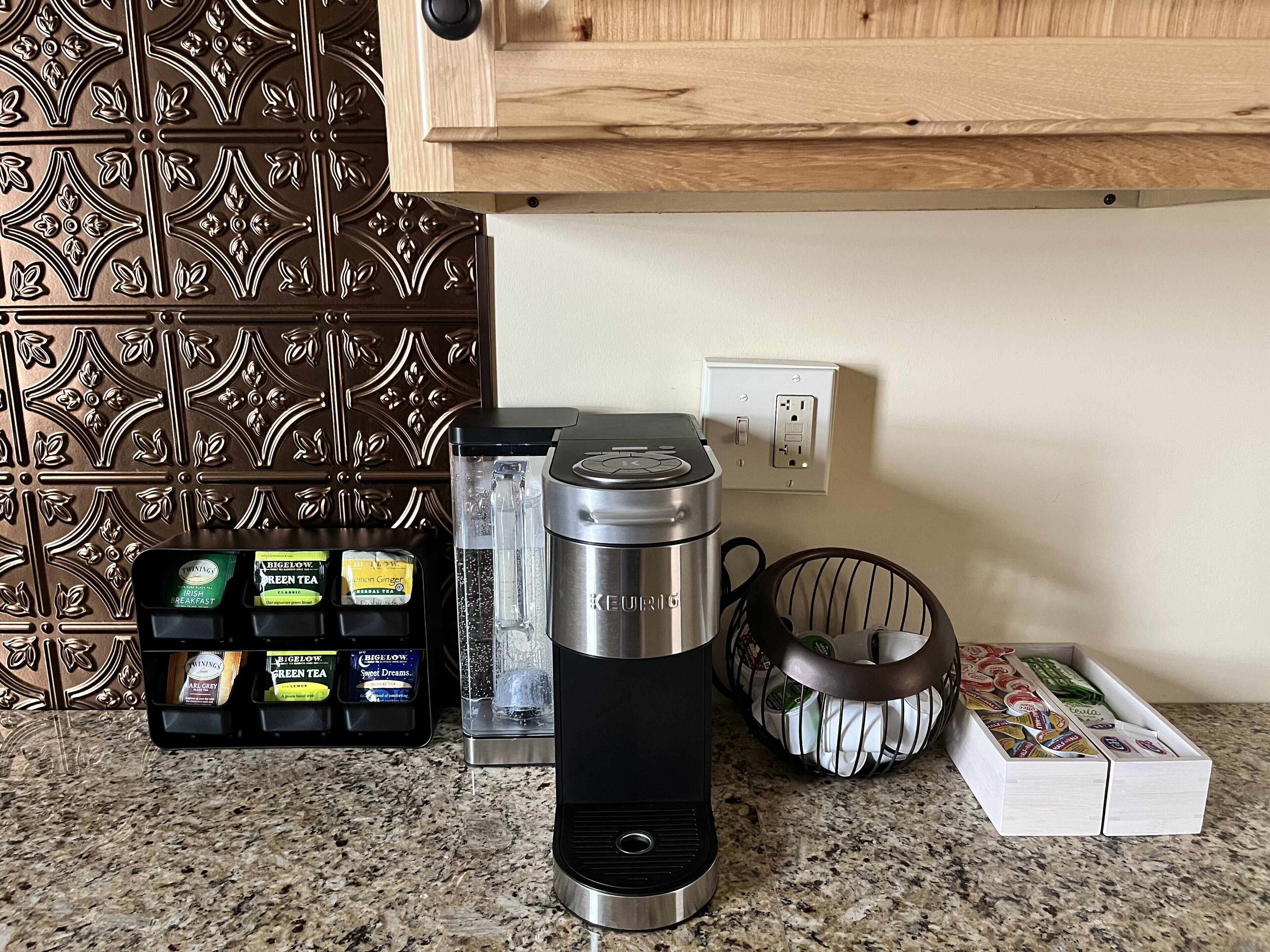 Una cocina con una cafetera Keurig, dispensador de agua, un organizador para cápsulas de café y una cesta llena de cápsulas de café.