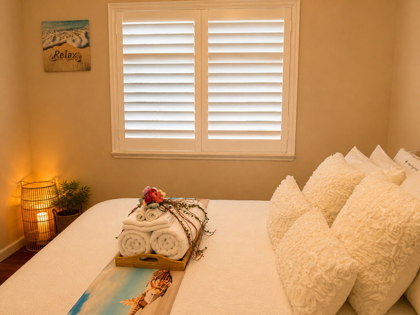 A bedroom with a bed, pillows, a tray with towels, a window with blinds, a wall decoration, and a small plant.