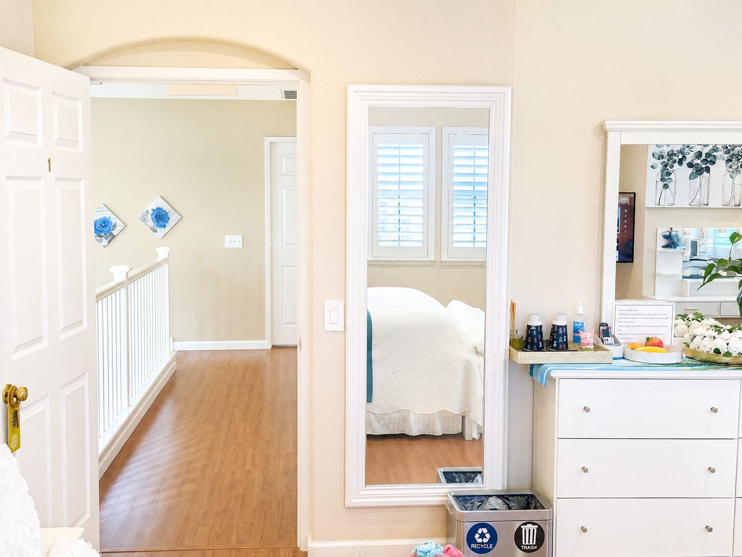 A hotel room with a bed, a mirror, a dresser, and a trash bin.