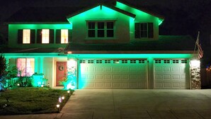 Uma casa de dois andares com garagem, iluminada por luzes verdes e uma bandeira americana à direita.
