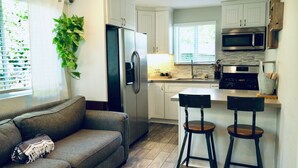 Una cocina moderna con armarios blancos, un refrigerador de acero inoxidable, un microondas y una estufa. Hay un taburete de bar y un sofá en la sala.
