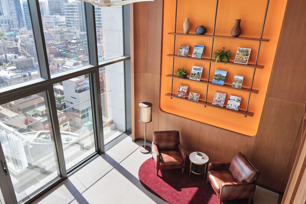A high-rise interior with a city view, a red carpeted area, leather seating, a round table, and a wall-mounted display of books and plants.