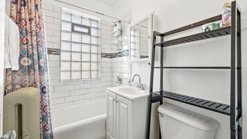 Une salle de bain avec une baignoire, un lavabo blanc, des toilettes, une douche et une fenêtre avec une grille en métal.