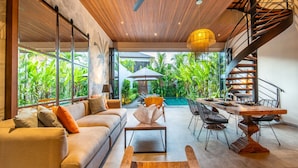 A modern living room with a sofa, coffee table, and dining table, a spiral staircase, and a view of lush greenery outside. - Private Pool • Garden Views • Two King Bedrooms (Canggu)