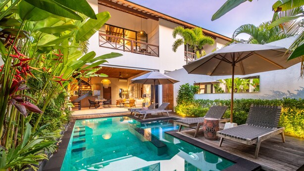 A swimming pool with lounge chairs and umbrellas, surrounded by lush greenery and a modern house with a balcony. - Private Pool • Garden Views • Two King Bedrooms (Canggu)