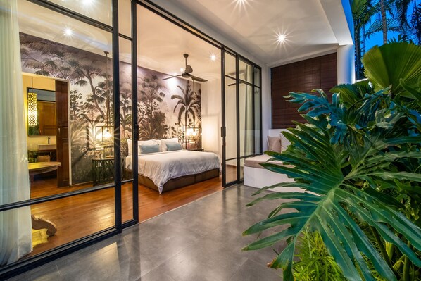 A modern bedroom with a large bed, a ceiling fan, and a view through glass doors to a bathroom.