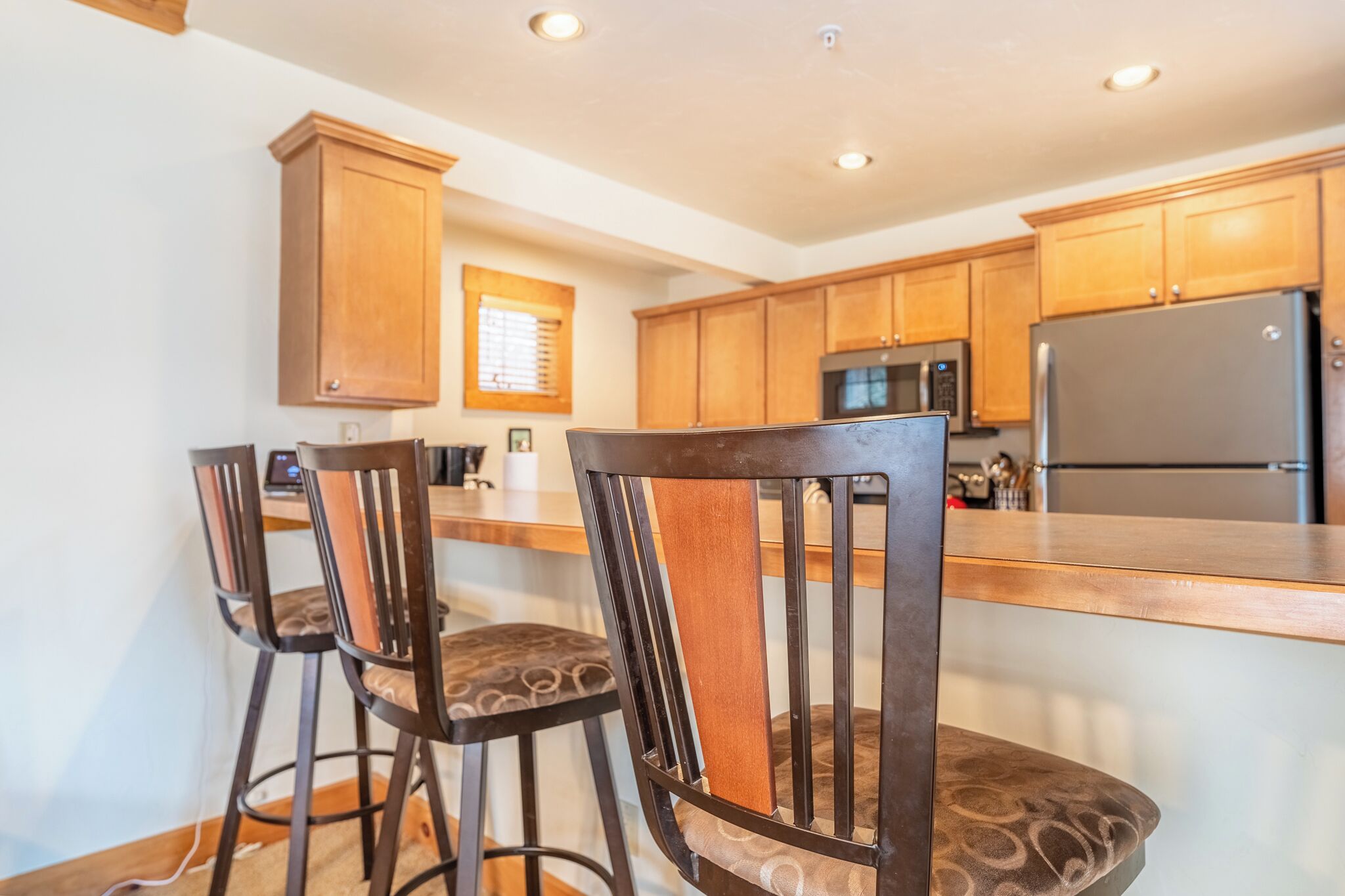 A kitchen with wooden cabinets, a stainless steel refrigerator, and two bar stools with patterned cushions.