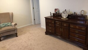 Ein Zimmer mit beigem Teppich, einem braunen Sessel mit gemustertem Kissen und einer Holzkommode mit mehreren Schubladen und Dekorationselementen.