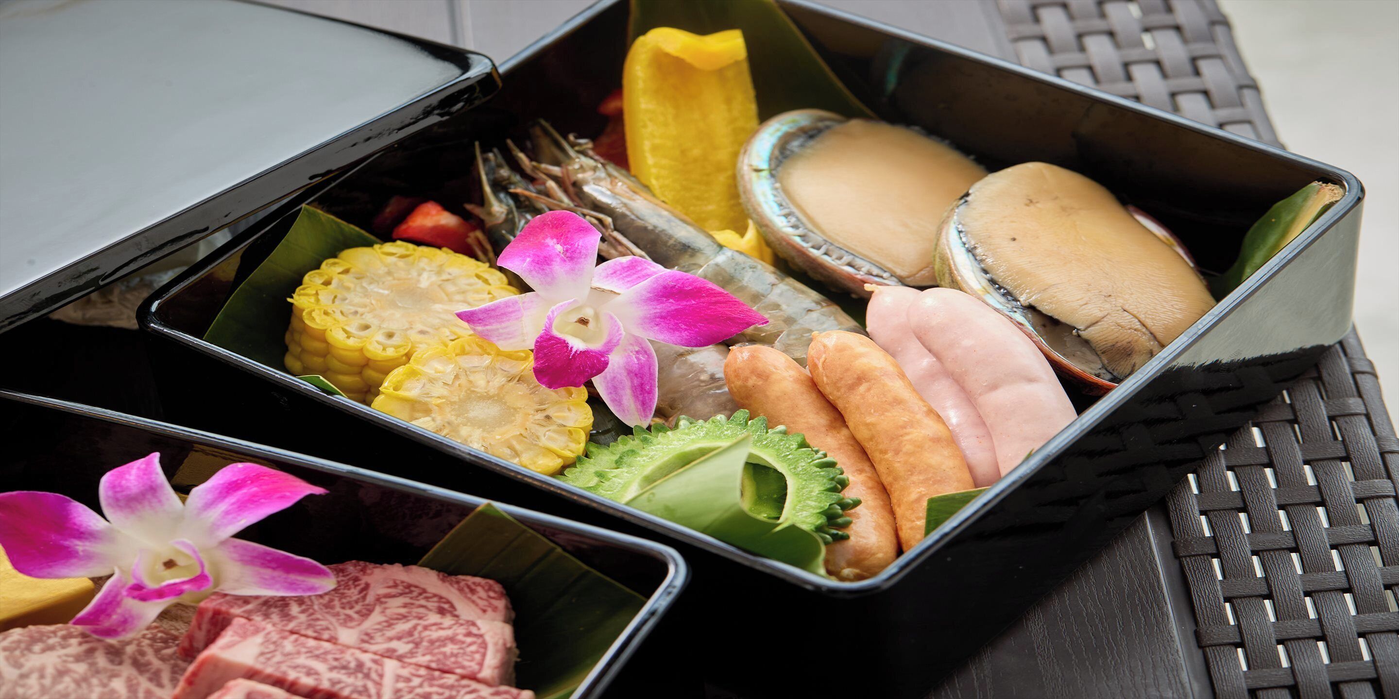 A tray with various food items including corn, shrimp, and a bun, arranged on a woven mat.