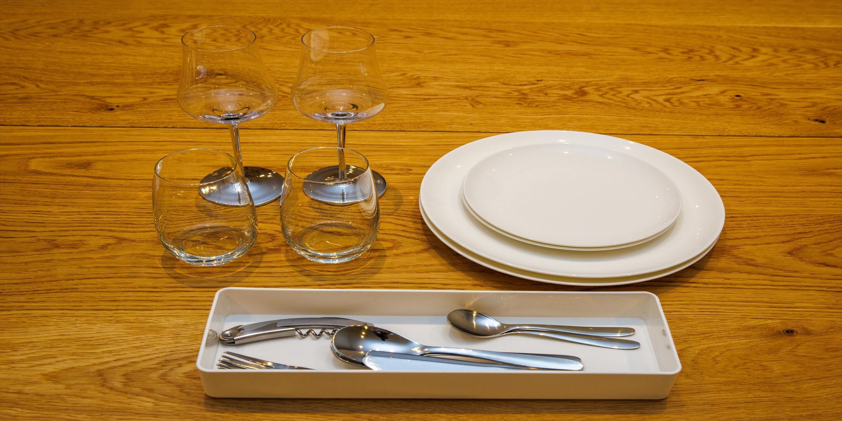 A wooden table with four wine glasses, a stack of white plates, and a rectangular tray with cutlery.