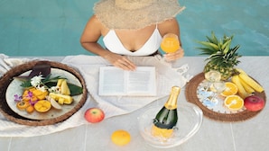 A person by a pool with a book, fruit, and drinks on a table. - ADAN RESORT Sky Villa luxury Suite (Motobu)