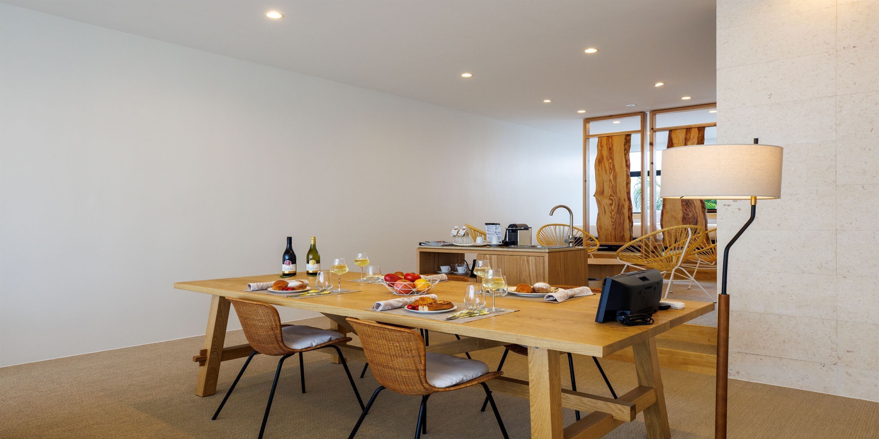 Un coin repas avec une table en bois, des chaises en osier et une lampe. On peut voir des bouteilles, des verres et de la nourriture sur la table.