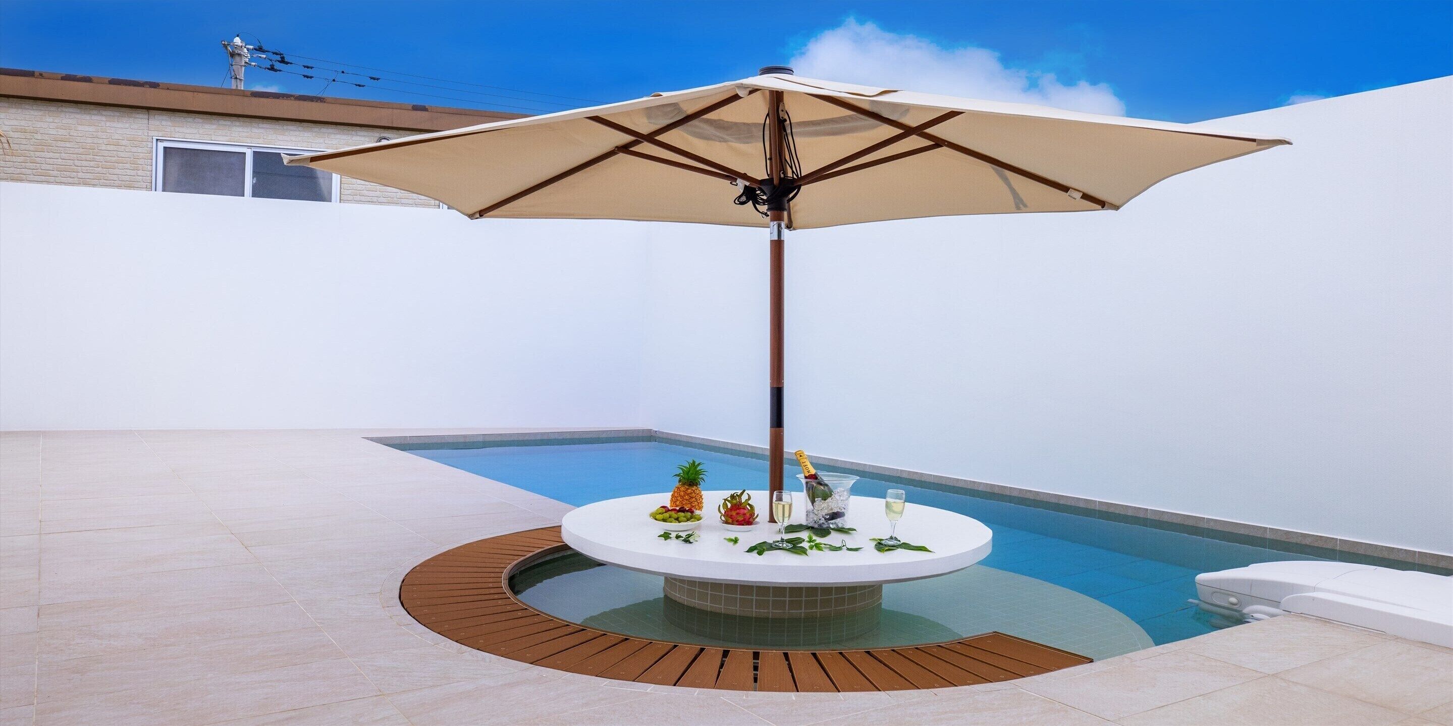 A poolside area with a round table, a large umbrella, and a bottle of champagne.
