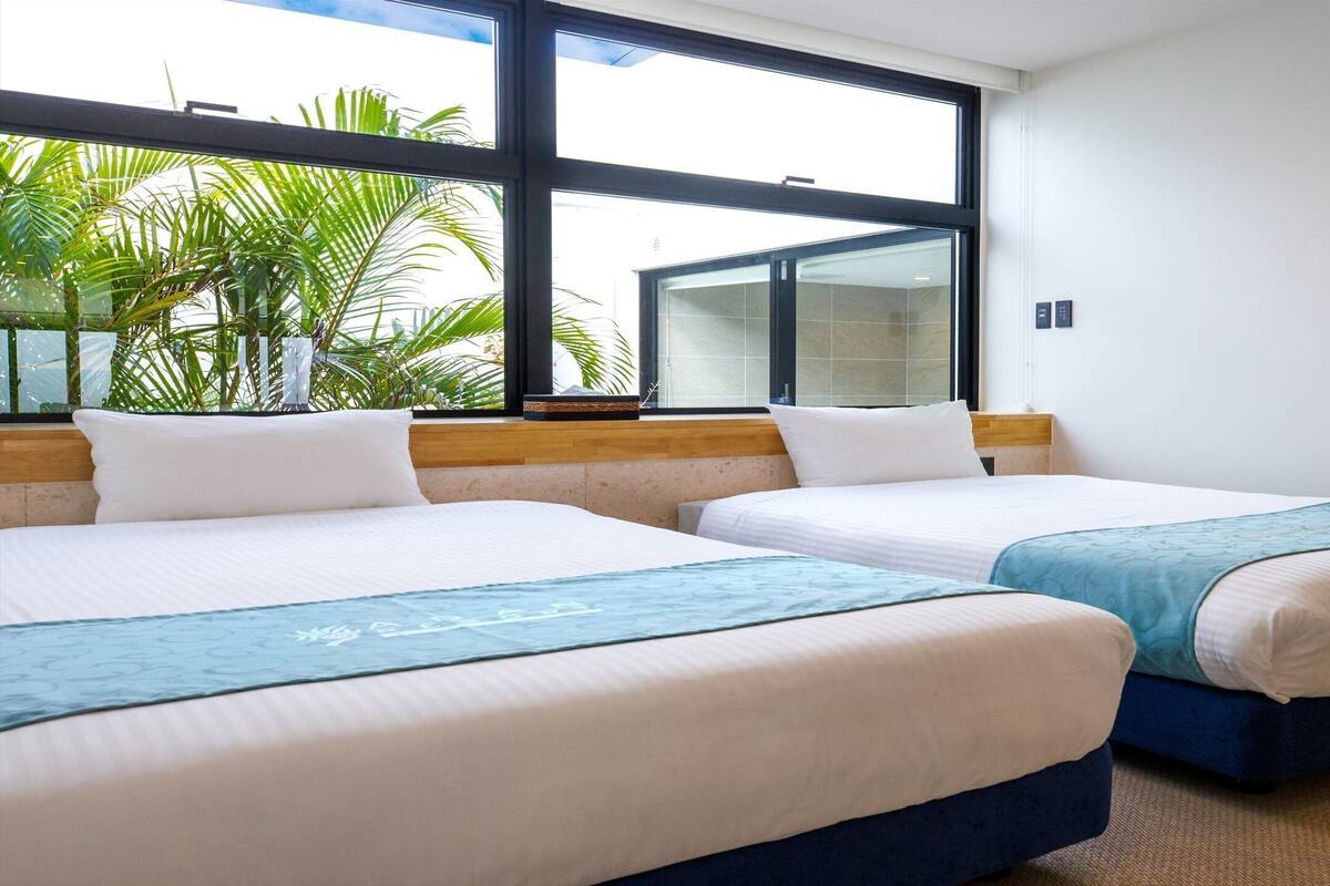 Ein Hotelzimmer mit zwei Betten, großen Fenstern und Blick auf Palmen.