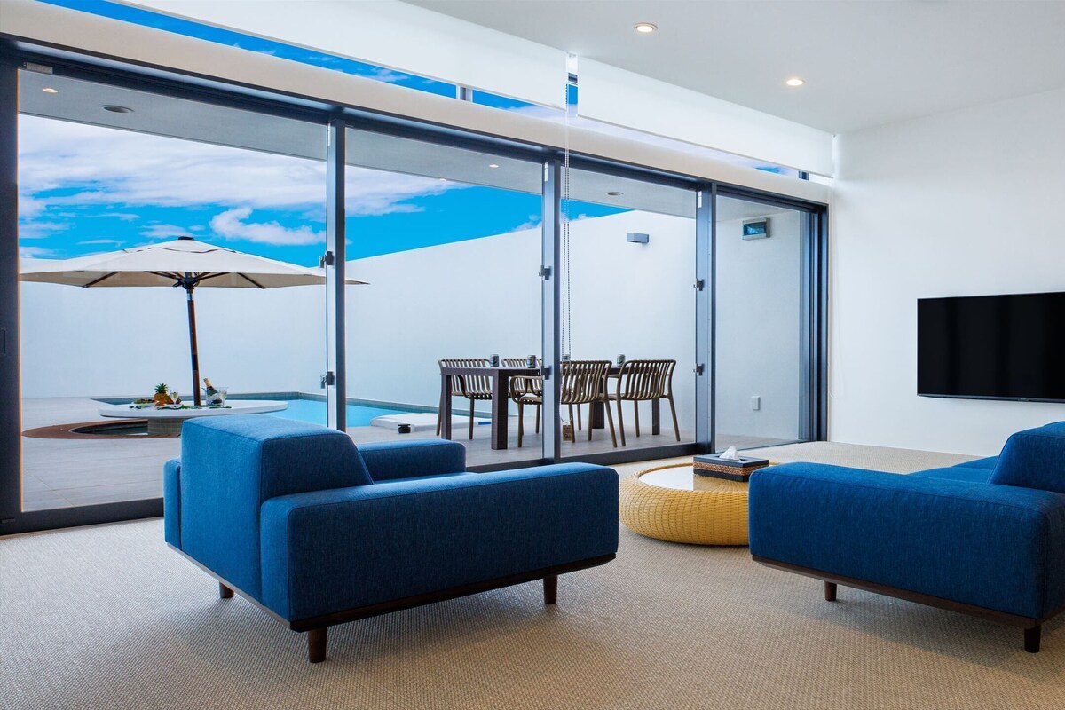 A modern living room with two blue sofas, a flat-screen TV, and a glass wall overlooking a pool area.