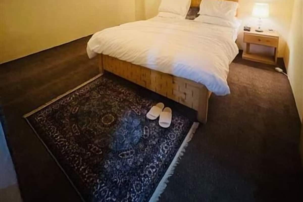 A bedroom with a wooden ceiling, a bed with white bedding, a nightstand with a lamp, and a patterned rug.