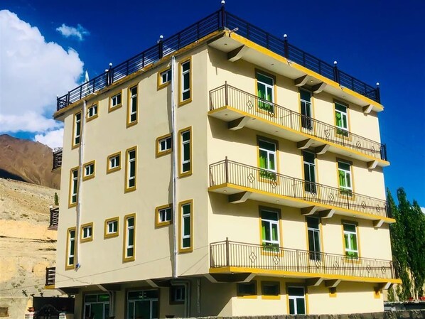 Un edificio de varios pisos con balcones, un techo plano y montañas al fondo.