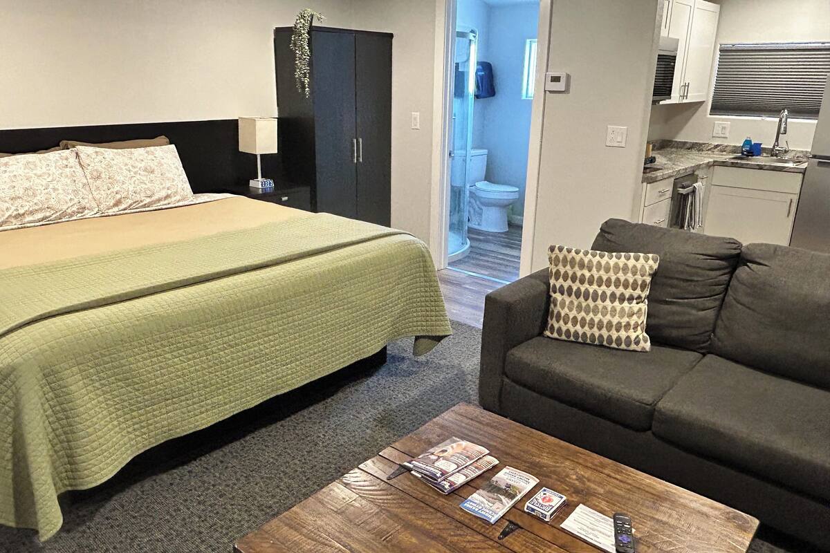 A hotel room with a bed, a sofa, a wooden coffee table with magazines, a nightstand, a lamp, a wardrobe, and a view into the bathroom.
