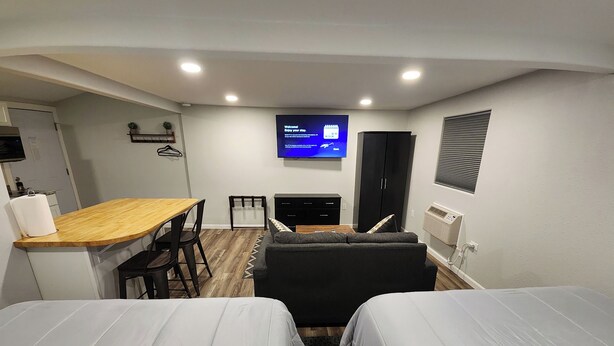 A compact hotel room with a kitchenette, a flat-screen TV mounted on the wall, a sofa, and two single beds.