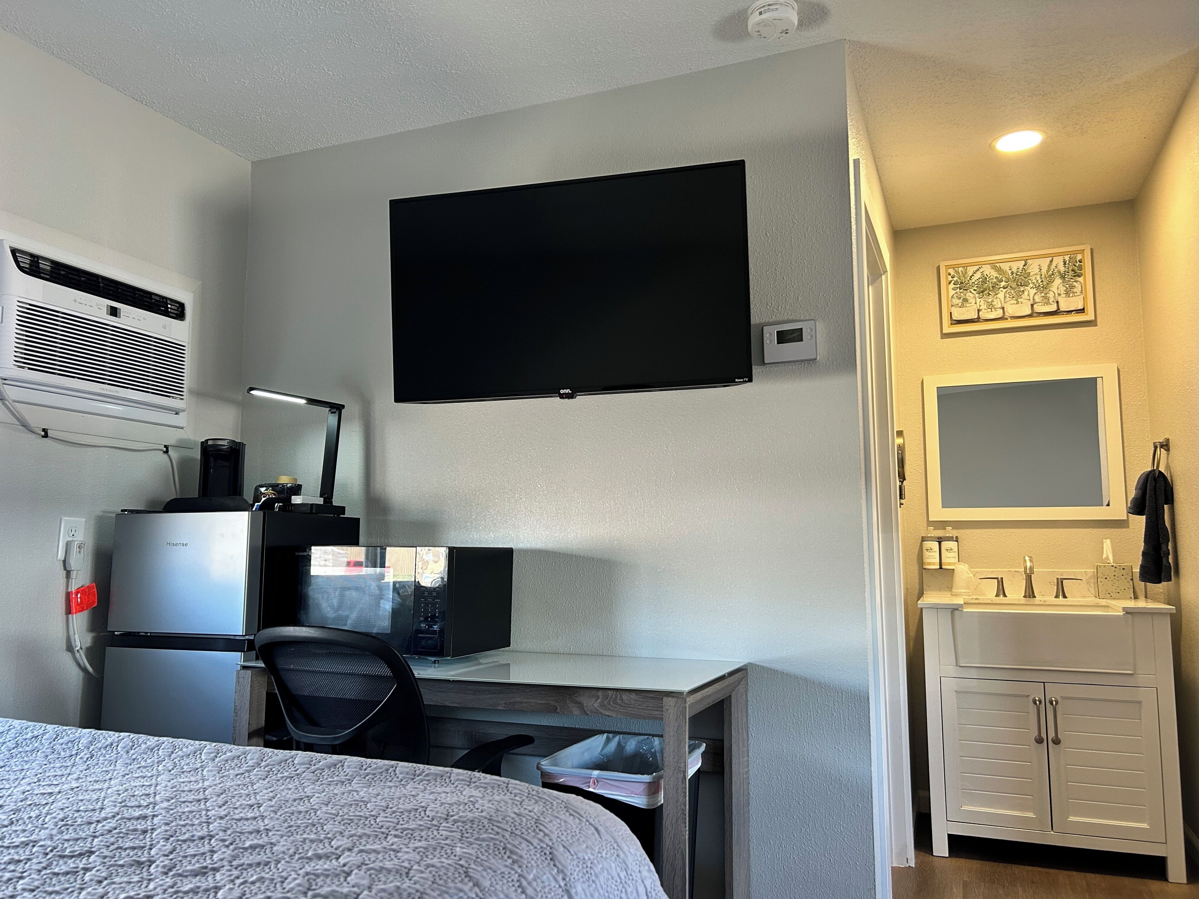 A hotel room with a bed, a desk with a computer, a flat-screen TV, a microwave, a coffee maker, a heater, a sink with a mirror, and a cabinet.