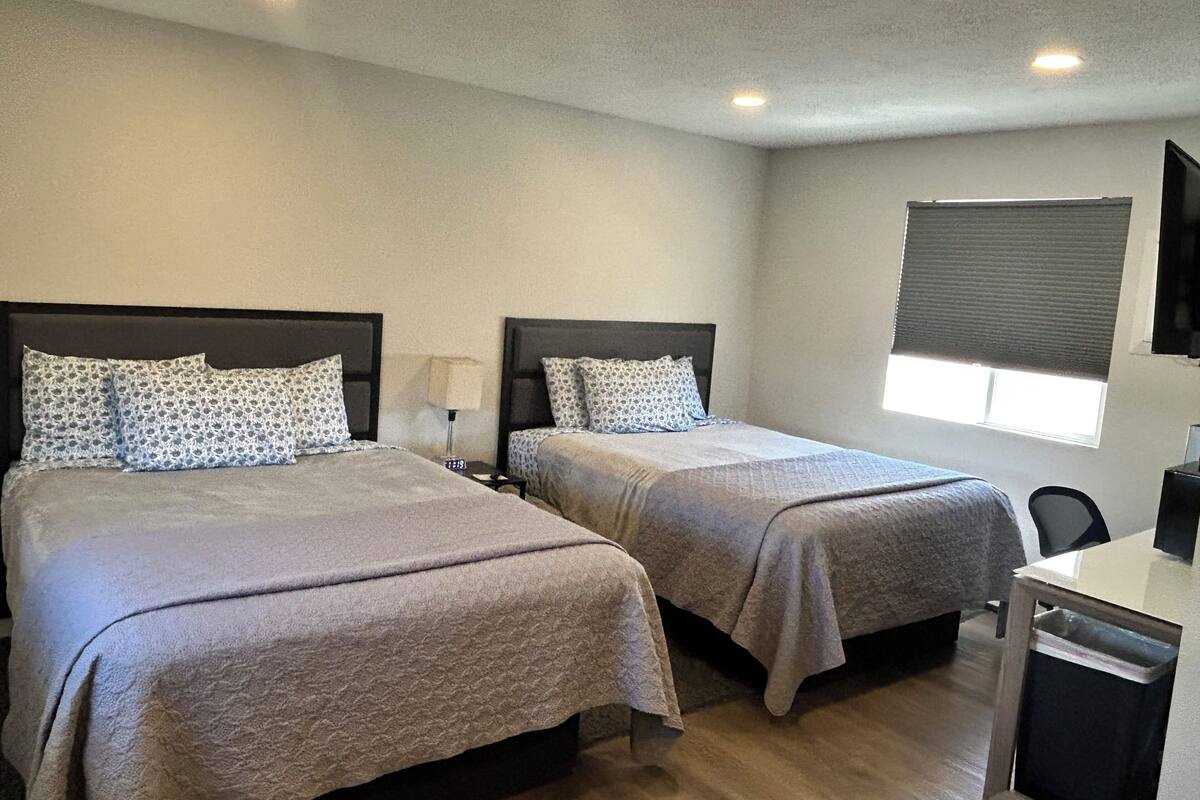 A hotel room with two beds, a desk, a chair, a TV, and a window with blinds.