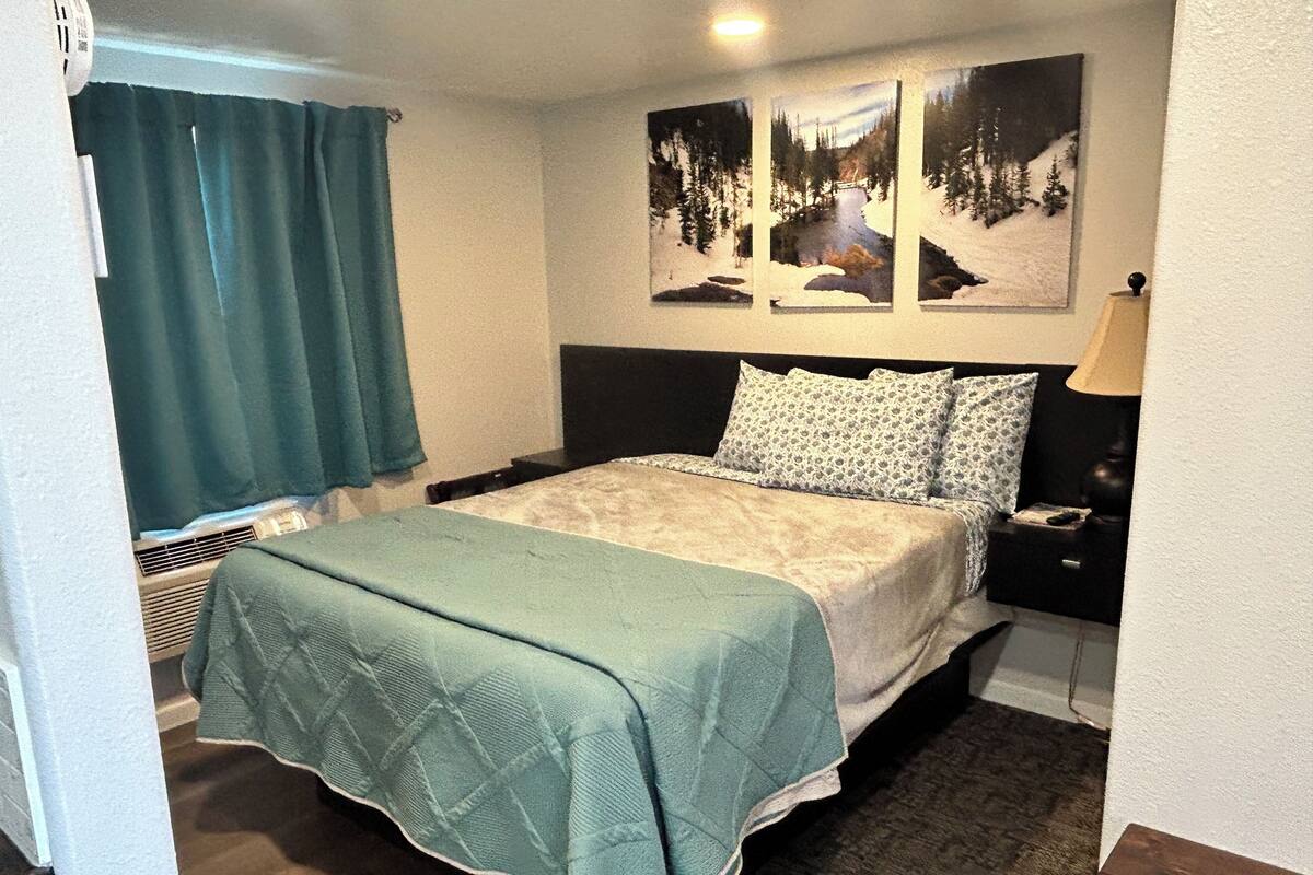 A bedroom with a bed, a nightstand, a lamp, a window with curtains, and a painting on the wall.