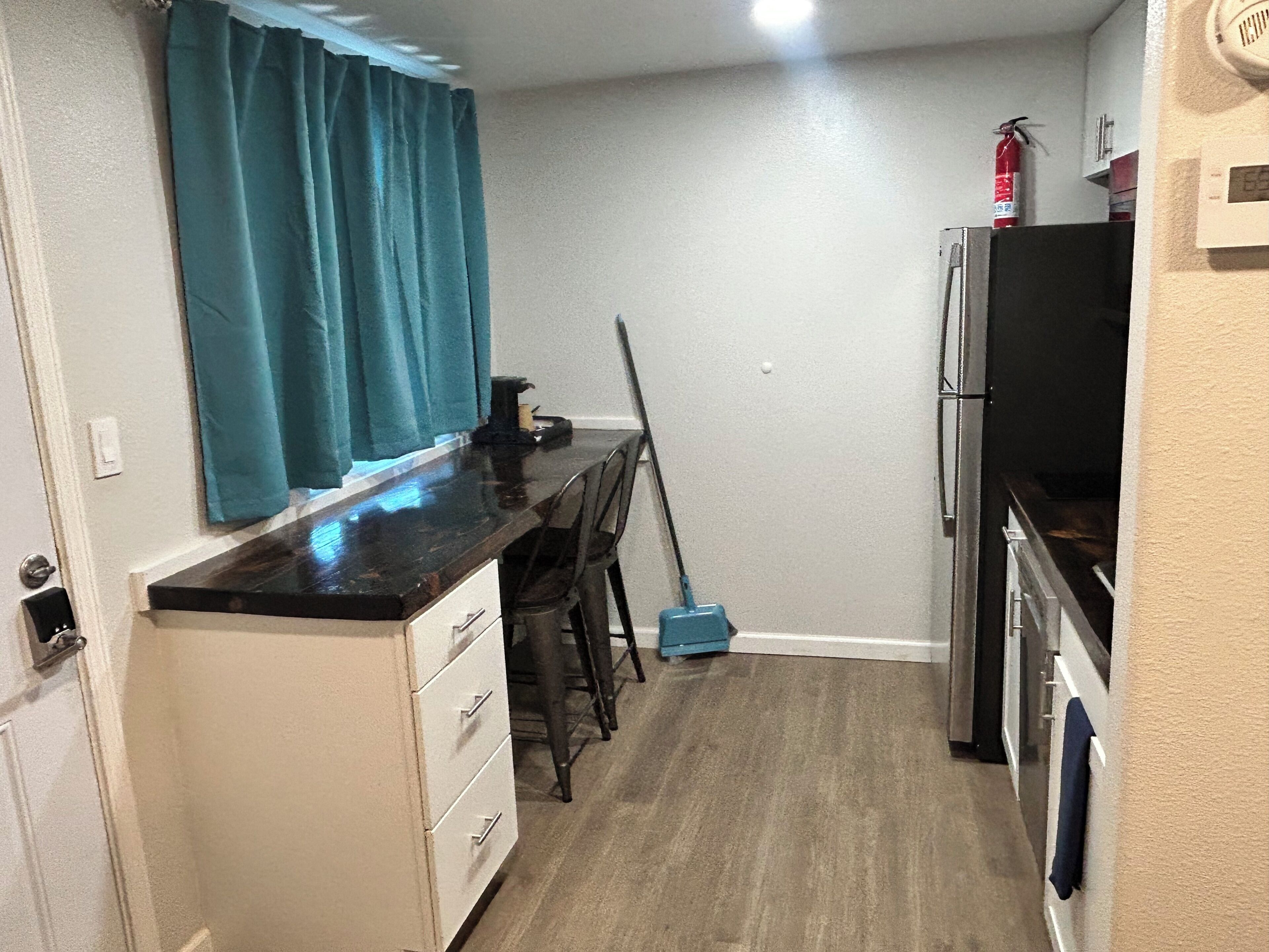 A compact kitchen with a countertop, a broom, a refrigerator, and a wall-mounted fire extinguisher.