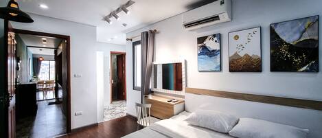 A modern hotel room with a bed, desk, and chair. There are three paintings on the wall and an air conditioning unit above the desk.