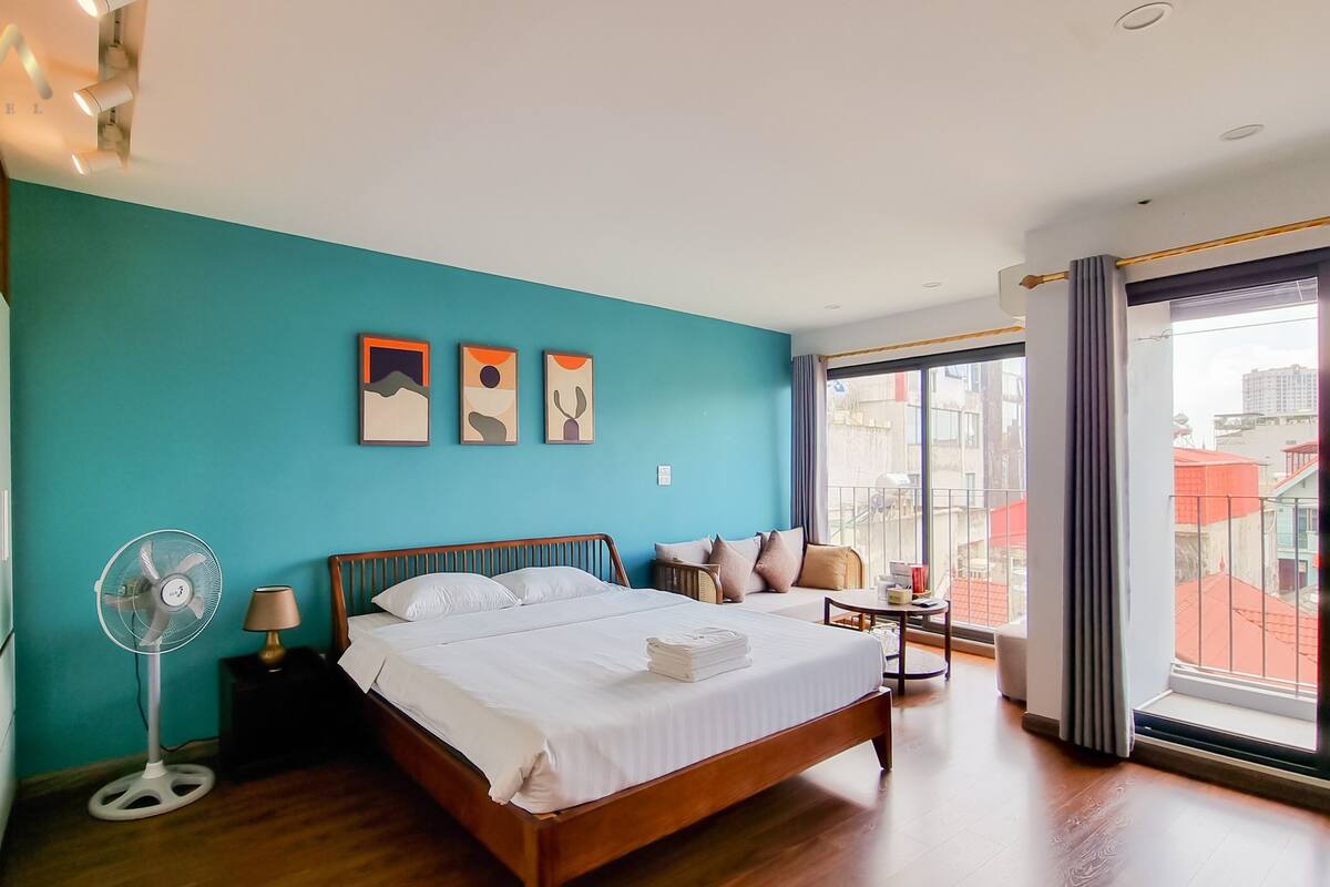 Une chambre d’hôtel moderne avec un mur d’accent de couleur turquoise, un lit en bois, un ventilateur sur pied et une vue sur la ville grâce à de grandes fenêtres.
