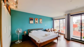 A modern hotel room with a teal accent wall, a wooden bed, a standing fan, and a view of the city through large windows.