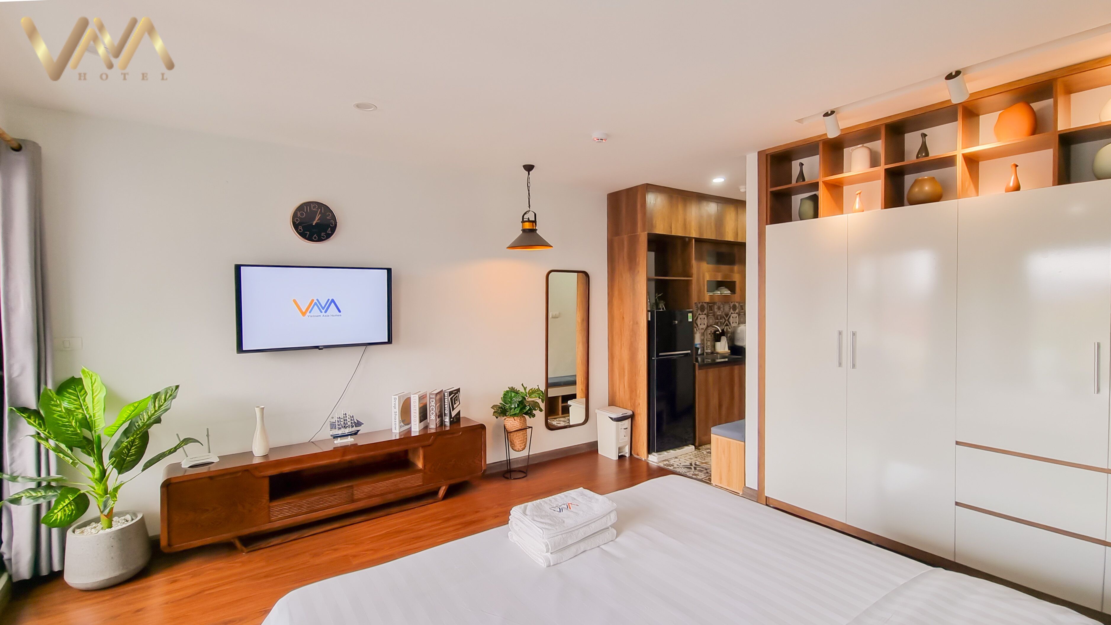 A modern hotel room with a bed, a TV mounted on the wall, a wooden wardrobe, and a small kitchenette.