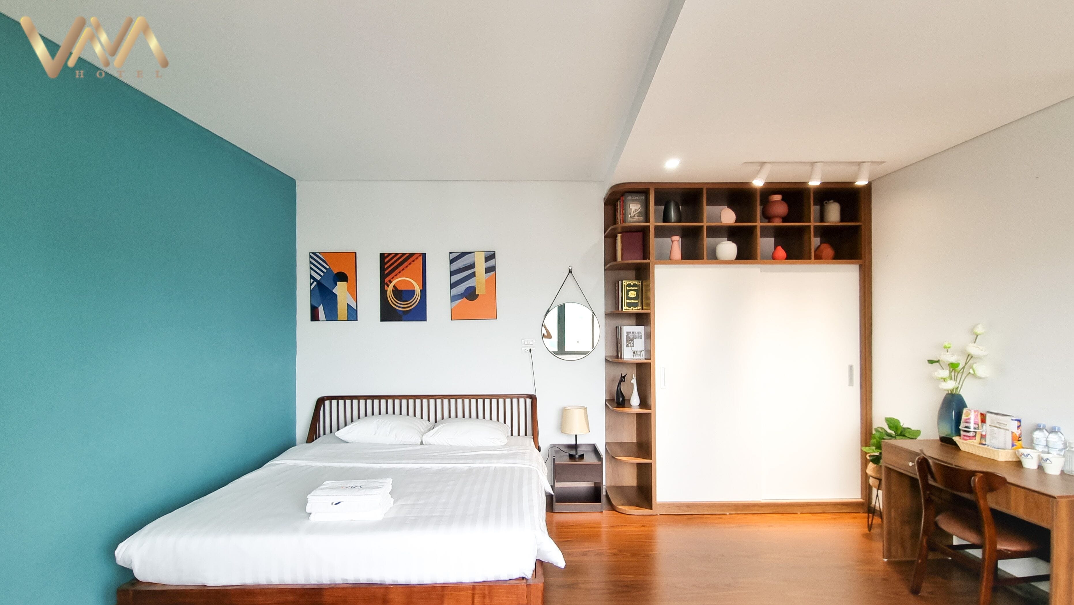 A modern hotel room with a bed, bedside table, lamp, and a bookshelf with decorative items.