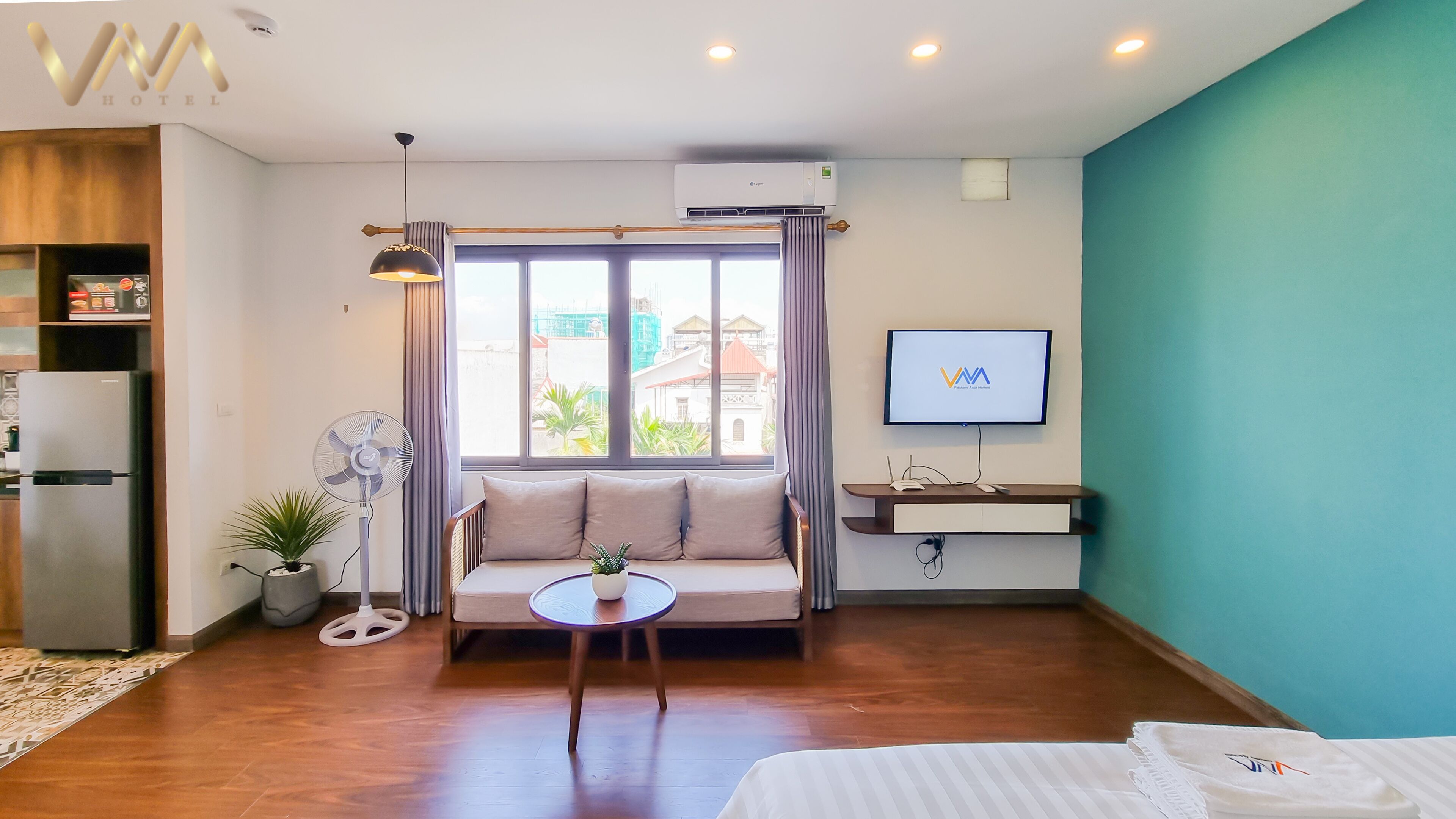 A hotel room with a bed, a sofa, a small table, a TV, an air conditioner, and a window with a view of buildings.