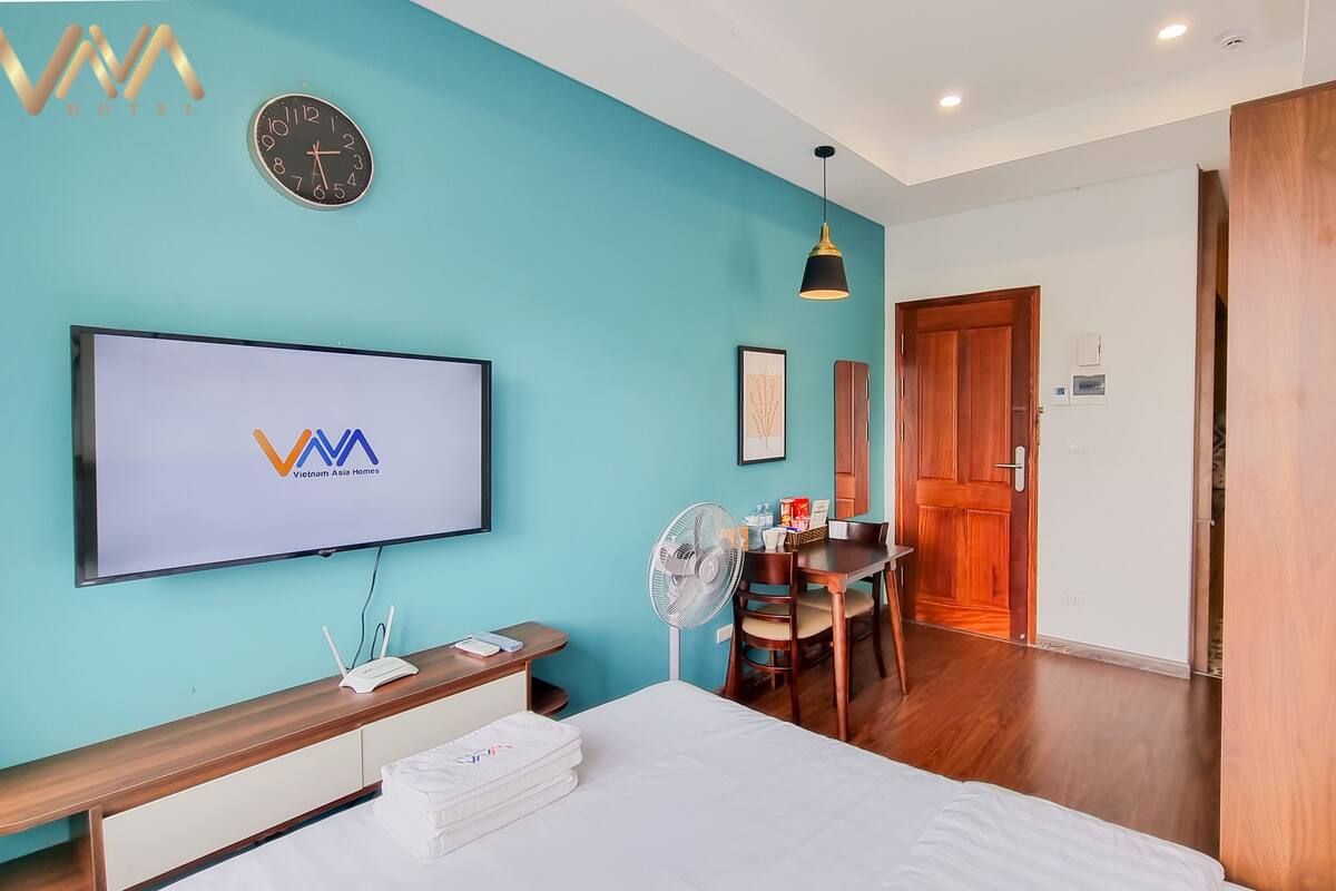 Une chambre d’hôtel moderne avec un mur de couleur turquoise, un téléviseur à écran plat affichant le logo de l’hôtel, un bureau en bois avec une chaise, une table de chevet avec une lampe et une grande armoire.