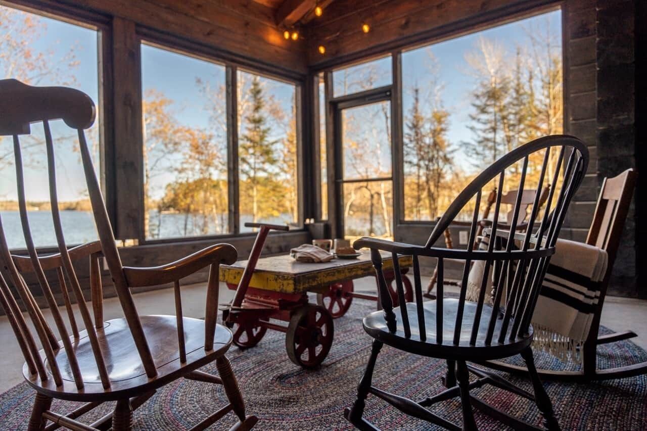 Un coin salon accueillant avec une table et des chaises en bois, de grandes fenêtres offrant une vue sur les arbres et un plan d’eau.