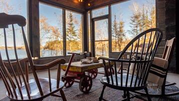 Un coin salon accueillant avec une table et des chaises en bois, de grandes fenêtres offrant une vue sur les arbres et un plan d’eau.