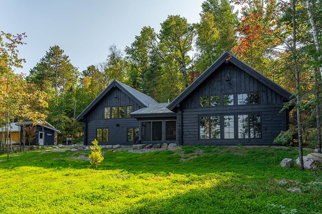 A two-story wooden cabin with a large window on the second floor, surrounded by a grassy yard and trees.