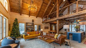 A spacious living area with a high ceiling, wooden beams, a Christmas tree, a sofa, a coffee table, and a kitchen area. - Northern Exposure Modern Log Cabin (Ely)