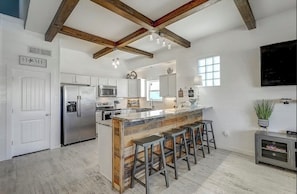 A modern kitchen with white cabinetry, a central island with bar stools, a stainless steel refrigerator, and a wooden ceiling beam structure.