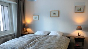 Ein Schlafzimmer mit einem großen Bett, zwei Nachttischen mit Lampen, einem Fenster mit Vorhängen und gerahmten Bildern an der Wand.