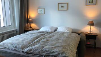 Ein Schlafzimmer mit einem großen Bett, zwei Nachttischen mit Lampen, einem Fenster mit Vorhängen und gerahmten Bildern an der Wand.