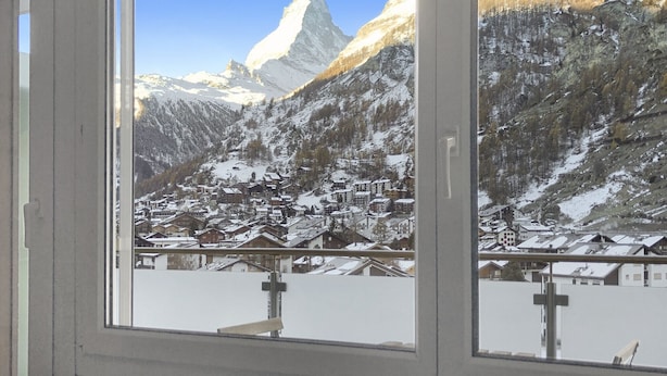 Blick aus dem Fenster auf ein Bergdorf mit schneebedeckten Häusern und einem markanten Berggipfel.