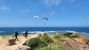 Zona costera con cielo despejado, costa rocosa y personas caminando por un sendero, con cometas volando por encima.