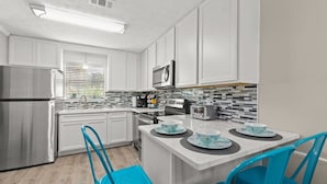 A modern kitchen with white cabinets, a stainless steel refrigerator, a microwave, and a dining area with blue chairs and a table set for four. - Absolute Beach ~ Pet-Friendly Townhome with Private Backyard (Panama City Beach)