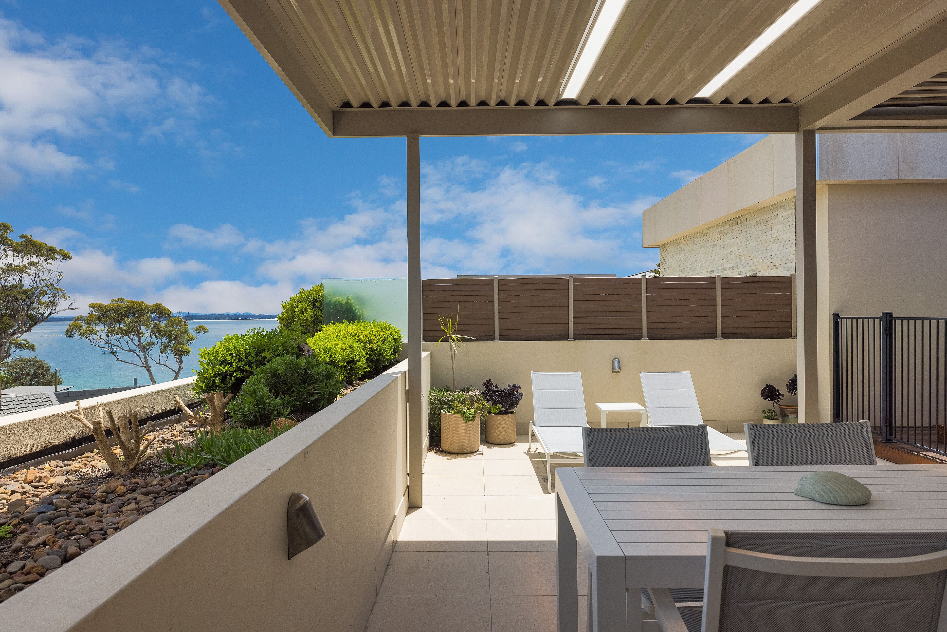 A covered outdoor patio with a view of the sea, a table with chairs, and potted plants.