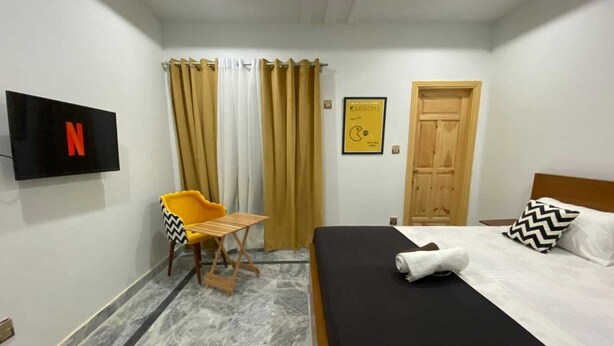 A hotel room with a bed, a TV mounted on the wall, a small table, and a chair. - Dastak Hotels  (Islamabad)